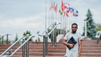 Black student posed with backpack and school items on yard of university, against flags of different countries.