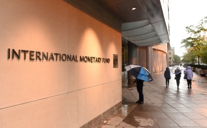 People with umbrellas walking past a building with a sign that says International Monetary Fund.