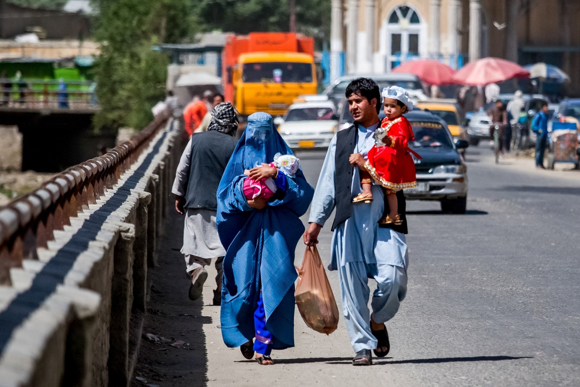 A man and woman, each carrying a small child, walk down a bridge next to a street.