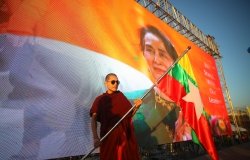 A man in red robes stands with a flag in front of a banner of Aung San Suu Kyi