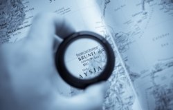 A hand holds a magnifying glass that is circling Brunei on a map.