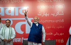 Narendra Modi stands in front of a red background, holding up his hand in a victory sign.